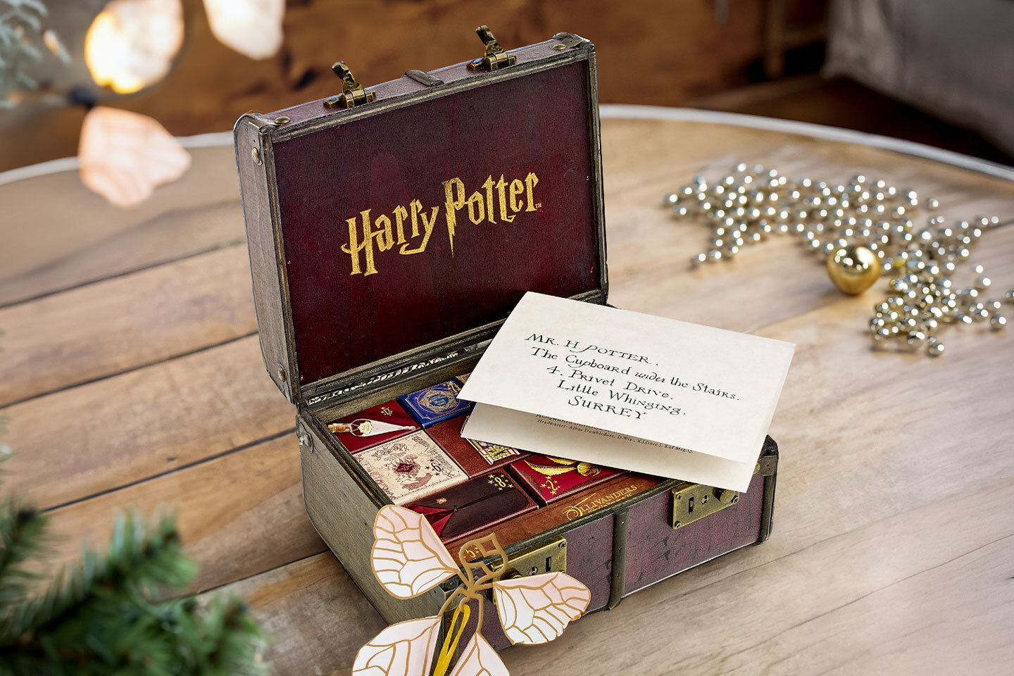 Geöffneter Harry Potter Adventskalender in Form eines nostalgischen Koffers mit kleinen Geschenkboxen und einem an Harry Potter adressierten Brief auf einem Holztisch. Im Hintergrund festliche Weihnachtsdekoration.